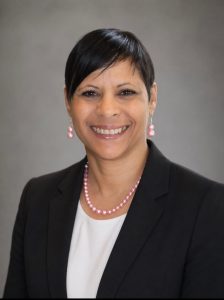 Headshot of a woman with dark, short hair. She's wearing a black blazer over a white blouse and has pink pearl jewelry on.