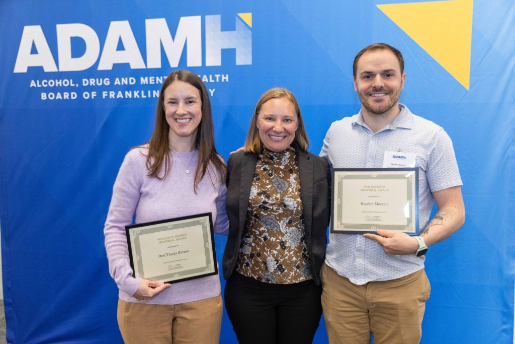 A group of three people stand smiling in front of an ADAMH banner, which stands for Alcohol, Drug and Mental Health Board of Franklin County. Two individuals are holding framed certificates.