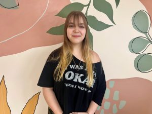 Image of a light skinned girl with bangs and long blonde hair smiling for a photo. She's wearing a baggy tshirt that's off the shoulder. Her background is a pastel painted wall of different shapes/shades of pink, tan and green.