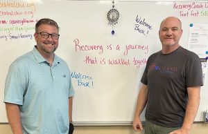 Two individuals standing in front of a whiteboard with motivational messages about recovery. The board includes phrases like "Recovery is a journey that is walked together" and "Welcome Back!" There are contact details for mental health resources. They are in a casual setting, dressed in relaxed attire.