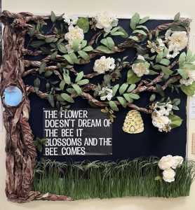 A decorative bulletin board features a tree with branches and leaves, white flowers, a beehive, and bees. A quote reads, "The flower doesn't dream of the bee. It blossoms and the bee comes."