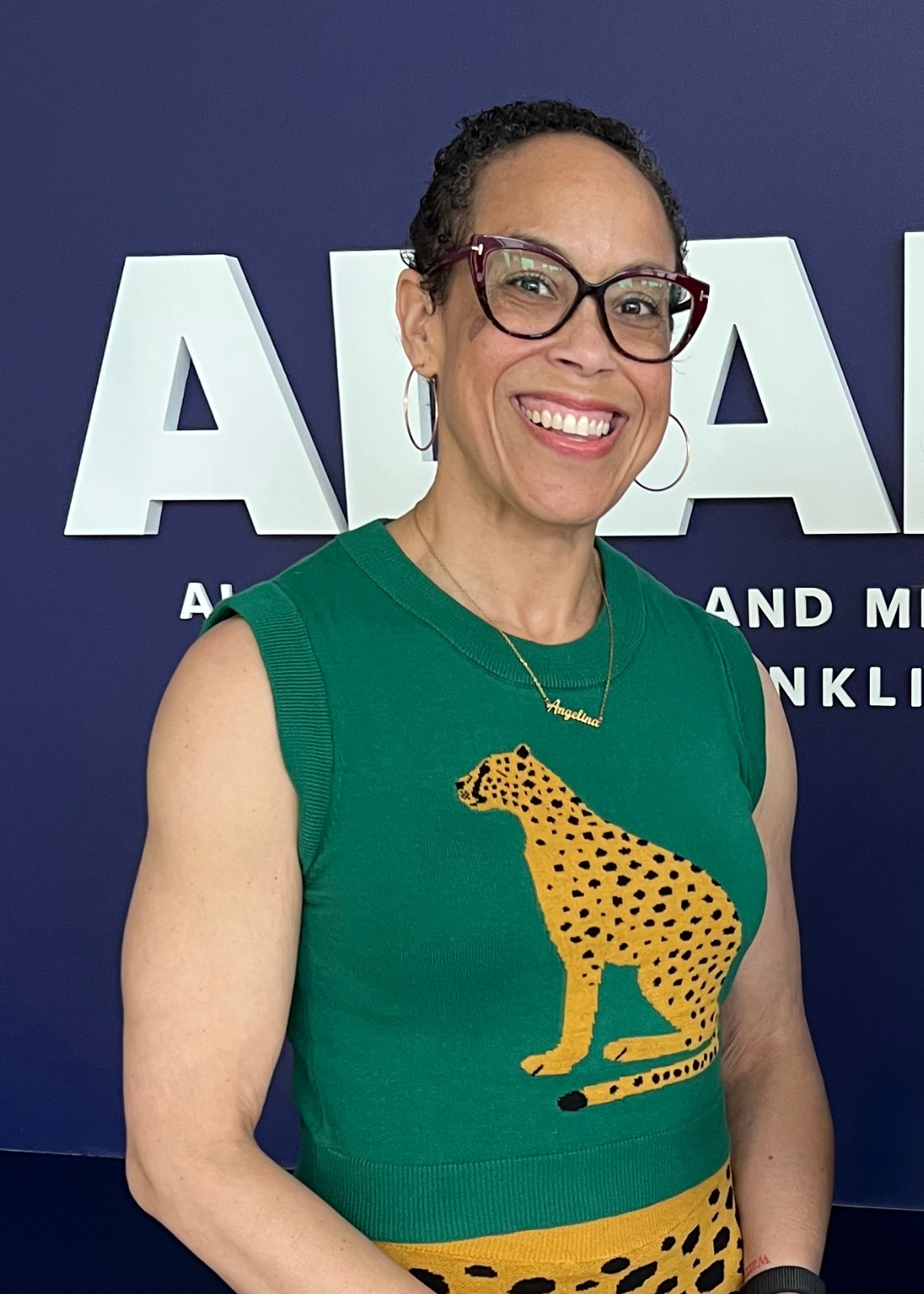 Woman with large rim glasses in a green sweater with a cheetah design on the front smiles for a photo.