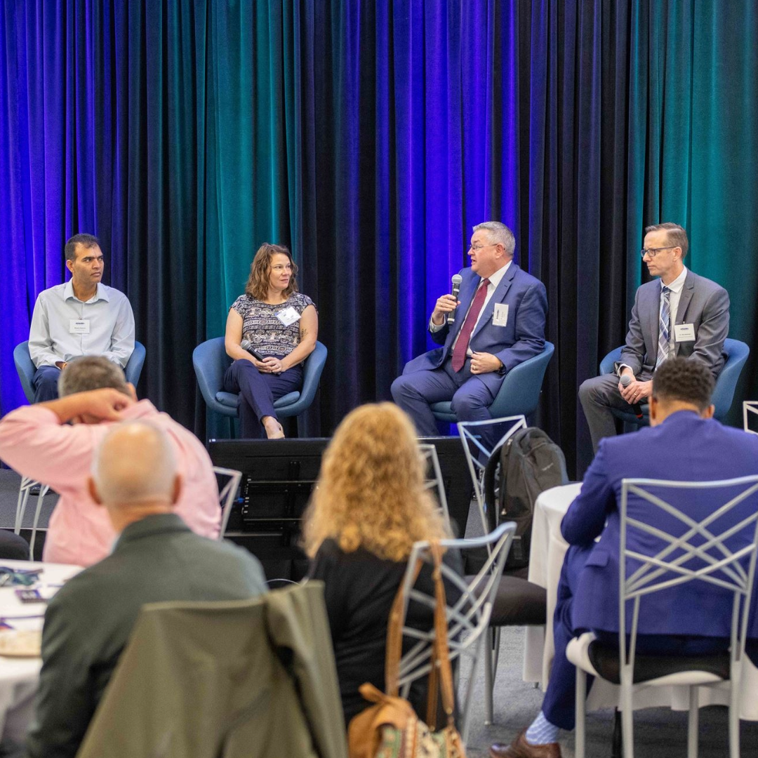 A panel of four individuals sits on a stage with microphones, engaged in discussion. The background features blue and green drapes. An audience is seated at round tables in the foreground, watching the panel. The setting appears to be a formal conference or event.