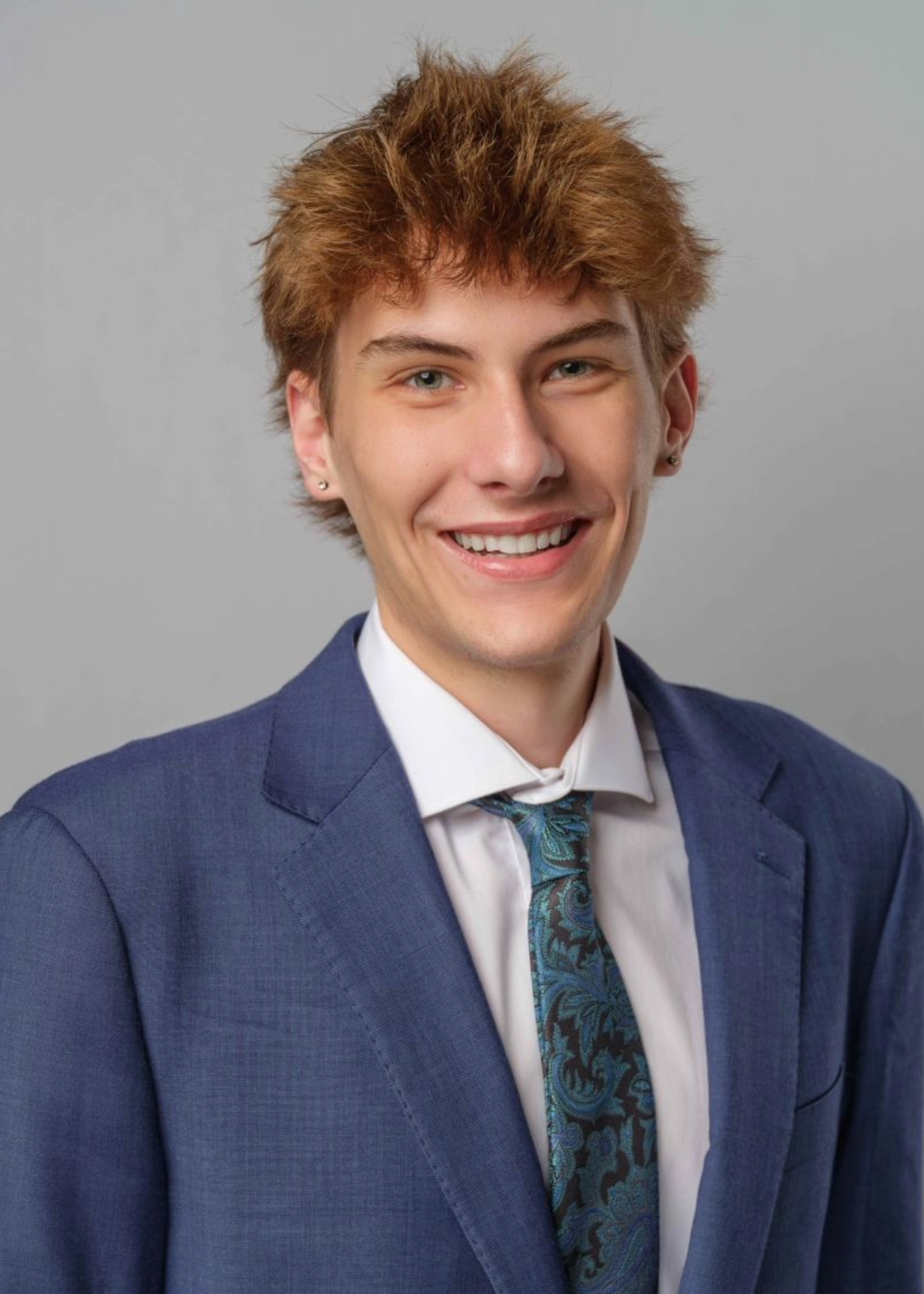 Light-skinned young man with light brown hair in a blue suit and turquoise tie smiling for a headshot