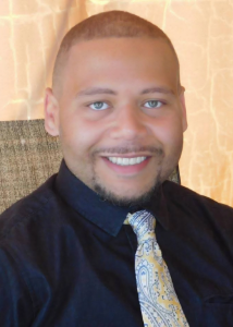 Headshot of a tan-skinned man wearing a black button up shirt and yellow patterned tie.
