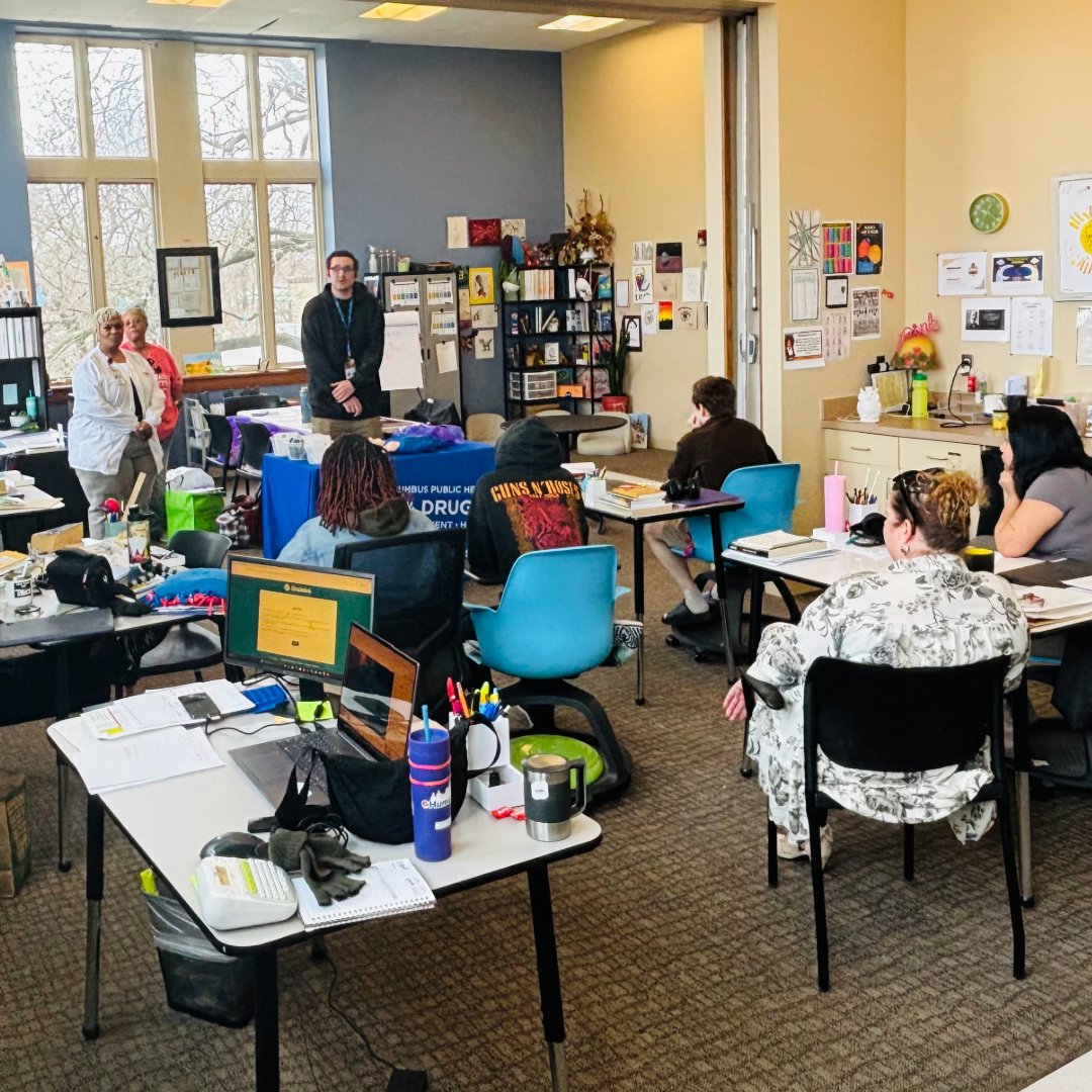 A group of people is gathered in a classroom setting, with desks and chairs arranged throughout the room. The participants, some seated and some standing, are engaged in activities. The room is well-lit with large windows, and walls are decorated with educational materials and artwork. There are various items on the tables, including a laptop displaying a website.