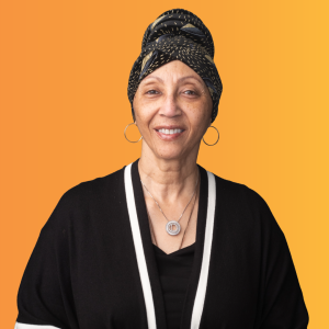 Woman in black with a black and gold accented headscarf with hoop earrings smiling at the camera against an orange background.