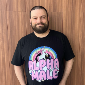 Headshot of man in black shirt with a unicorn and rainbow on it that says Alpha Male