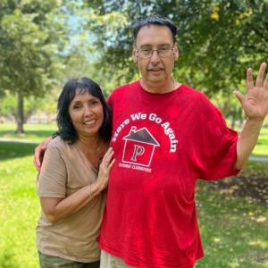 Man and woman pose for a photo in a park. The woman has dark hair and a tan shirt on while the man also has dark hair and a bright red t-shirt on.